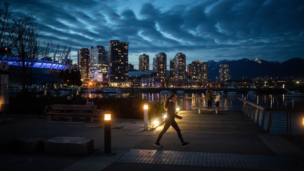 A woman walks on the seawall along False Creek as downtown condo towers are seen across the water, in Vancouver, on Friday, April 2, 2021. (Darryl Dyck / THE CANADIAN PRESS)